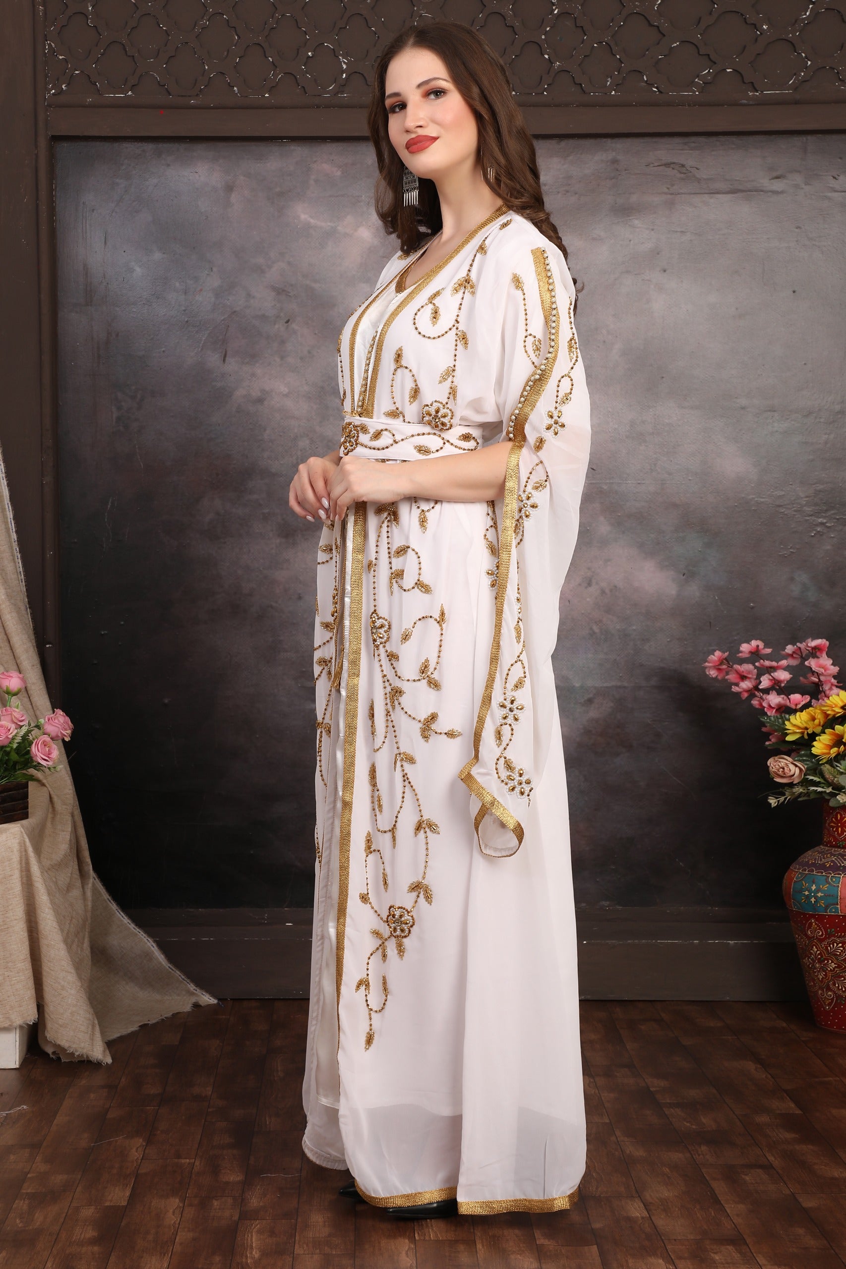Woman in a white dress with gold embroidery standing against a textured wall.