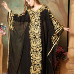 A woman modeling a black and gold embellished abaya dress with long sleeves and a flowing drape, posed in front of a floral backdrop.
