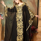 A woman modeling a black and gold embellished abaya dress with long sleeves and a flowing drape, posed in front of a floral backdrop.