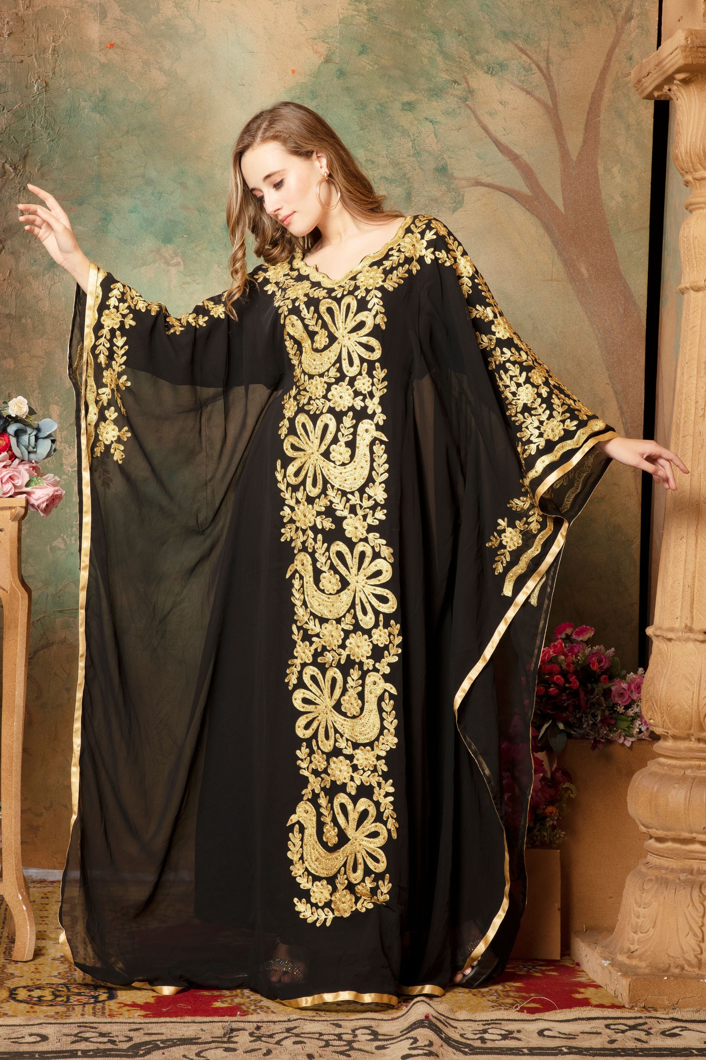 A woman modeling a black and gold embellished abaya dress with long sleeves and a flowing drape, posed in front of a floral backdrop.