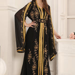 A woman wearing a black and gold Moroccan caftan with floral patterns and long sleeves, standing in a room with classic decor.