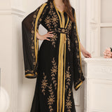A woman wearing a black and gold Moroccan caftan with floral patterns and long sleeves, standing in a room with classic decor.