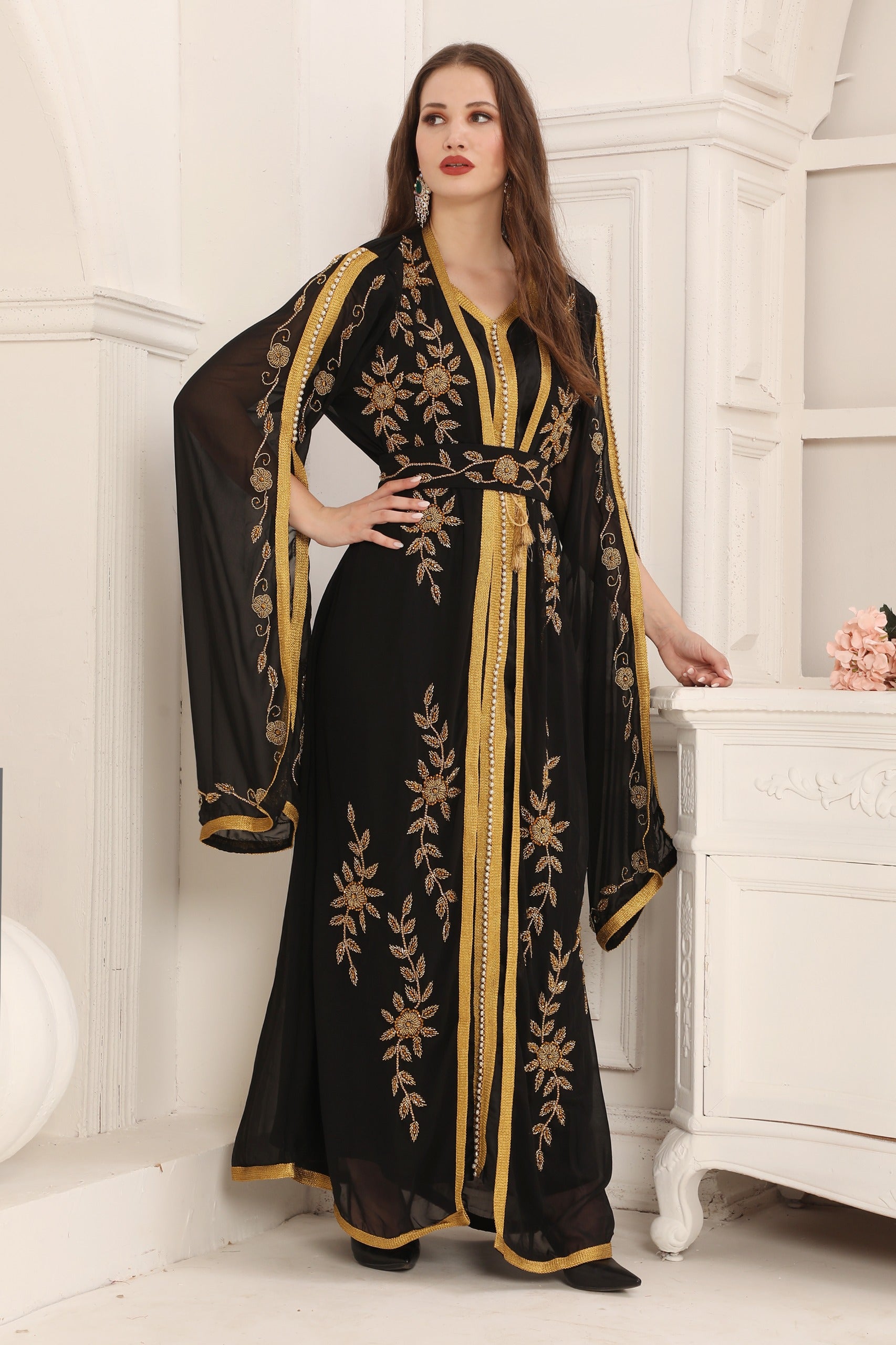 A woman wearing a black and gold Moroccan caftan with floral patterns and long sleeves, standing in a room with classic decor.