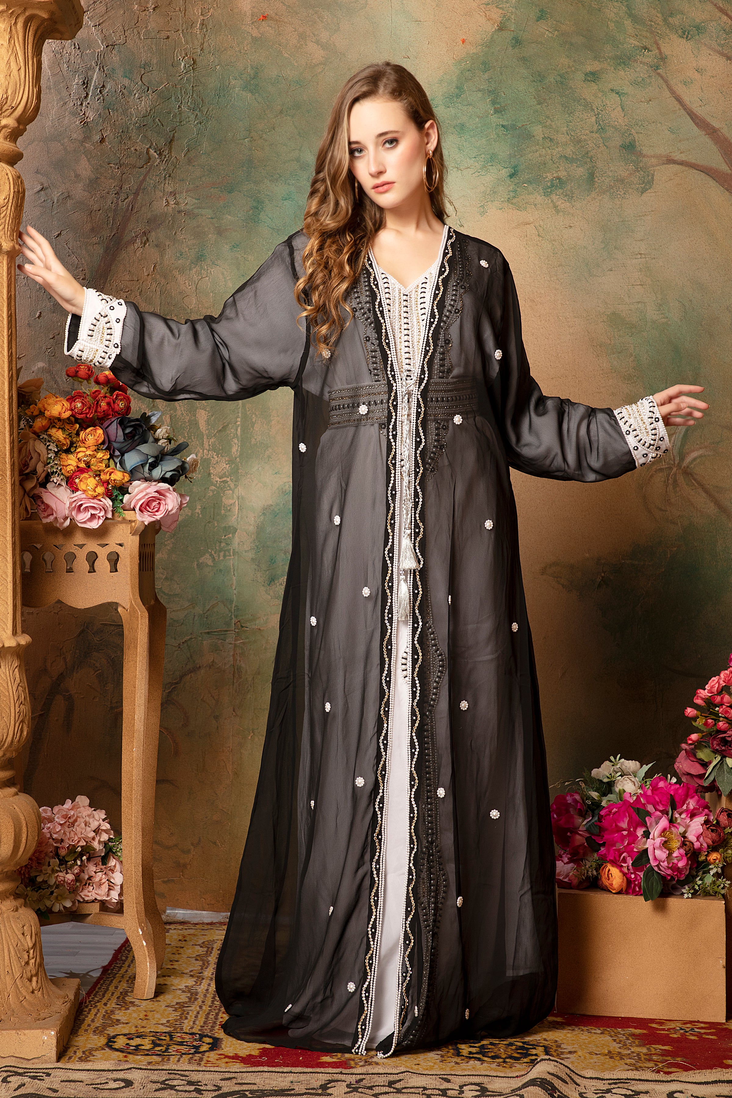 A woman modeling a black caftan with silver bead embellishments and a belt, standing in a floral backdrop.