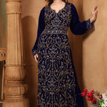 A woman wearing a navy blue embroidered gown with long sleeves, standing next to a column in a room with decorative flowers.