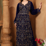 A woman wearing a navy blue embroidered gown with long sleeves, standing next to a column in a room with decorative flowers.