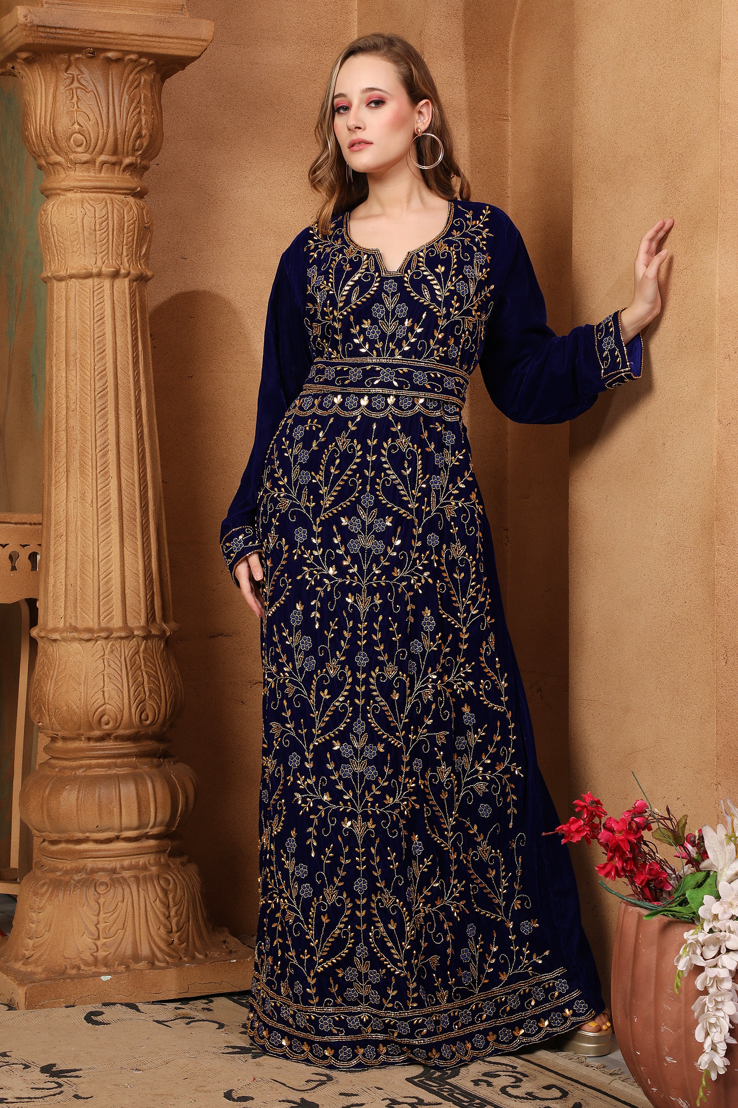 A woman wearing a navy blue embroidered gown with long sleeves, standing next to a column in a room with decorative flowers.