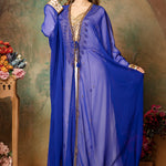 A woman wearing a blue caftan with golden bead embellishments, posing against a floral backdrop.