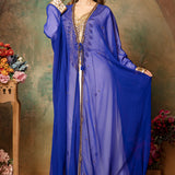 A woman wearing a blue caftan with golden bead embellishments, posing against a floral backdrop.