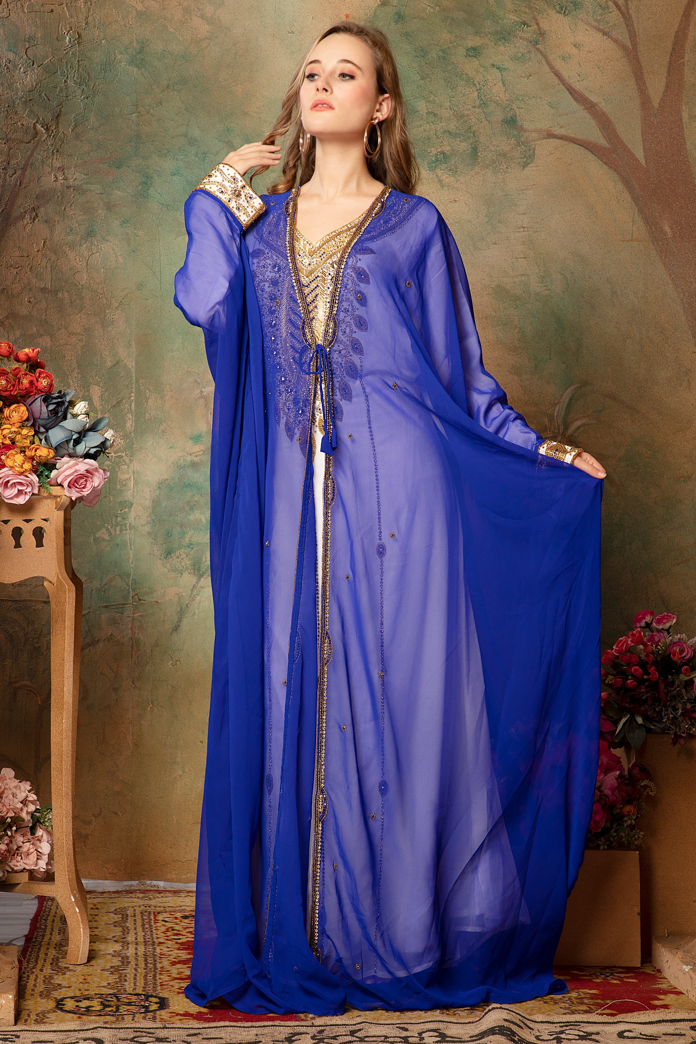 A woman wearing a blue caftan with golden bead embellishments, posing against a floral backdrop.
